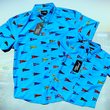 Load image into Gallery viewer, Full view of both the adult and youth 7-Strong "Pennant to Win It" shirt, which is sky blue with texture, featuring various triangular flags featuring baseball's prominent cities patterned throughout. The shirt is featured against abackground is sunset at a major league stadium.