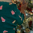 Load image into Gallery viewer, Male youth model sporting the 7-Strong "Oh, Christmas Treat" shirt - a green, christmas sweater-like background with columns of various white Christmas tree shaped cakes with red garland, some whole, some bitten. Model is putting a tree cake "ornament" on a tree.