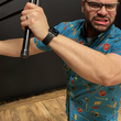 Load image into Gallery viewer, Medium view of male model swinging a steel chair at camera, wearing the 7-Strong "TLC OMG" shirt, a homage to wrestling objects featured all over the shirt including tables, ladders, chairs, ring bells, sledgehammers, brass knuckles, championships, guitars, stop signs, microphones amongst other items. The items are on a blue shirt with a faded steel cage background. The shirt is featured on a background of a wrestling ring with a standing ladder.