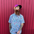 Load image into Gallery viewer, Medium shot of adult male in the 7-Strong Block Party adult button down, featuring various colored blocks of different arrangements inspired by a classic video game, cascading down the shirt against a grayish background featuring shadows of the same shapes. Model is standing against red slatted building wall.