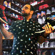 Load image into Gallery viewer, Male model wearing he adult short sleeve version of "Got 'Em!" The button down features various athletic fashion sneakers in an 8-bit format arranged all over a black shirt. Model is looking at two different pairs of choose trying to pick between them in a room full of other sneakers