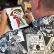 Load image into Gallery viewer, Over the shoulder of male model view of the adult 7-Strong "Cinco de Mayhem" button down shirt. Shirt features black and white posters featuring various created luchadors advertised for matches, all overlapping each other over a black shirt. Model is drawing one of the superstars featured on the shirt.