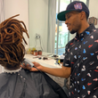 Load image into Gallery viewer, Male model wearing he adult short sleeve version of "Got 'Em!" The button down features various athletic fashion sneakers in an 8-bit format arranged all over a black shirt. Model is a barber shaping up the hairstyle of a patron.