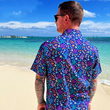 Load image into Gallery viewer, Medium close up, behind the back view of a male model of medium build wearing the 7-Strong "Coral of the Story" adult button down shirt. Color of the shirt is blue with vibrant purple, teal, orange sea life imagery - particularly fish and coral reef. The model is standing on the beach looking out towards the water wearing the shirt.