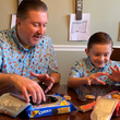 Load image into Gallery viewer, Father and son models wearing the 7-Strong "Cheat Day" adult button down featuring various snacks, treats, and fast food features on a light blue shirt. Both models are indulging in cookies and treats.
