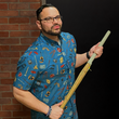 Load image into Gallery viewer, Full view male model holding a kendo stick against a brick wall,wearing the 7-Strong "TLC OMG" shirt, a homage to wrestling objects featured all over the shirt including tables, ladders, chairs, ring bells, sledgehammers, brass knuckles, championships, guitars, stop signs, microphones amongst other items. The items are on a blue shirt with a faded steel cage background. The shirt is featured on a background of a wrestling ring with a standing ladder.