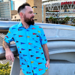 Load image into Gallery viewer, Medium shot view of a male model of the 7-Strong "Pennant to Win It" shirt, which is sky blue with texture, featuring various triangular flags featuring baseball's prominent cities patterned throughout. The shirt is being worn out front of Petco Park in San Diego.