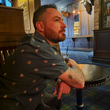 Load image into Gallery viewer, Male model wearing the 7-Strong "Straight Sippin'" Short Sleeve button down. The shirt is a deep green with black ripples as its background, while the foreground has various drinking glasses with various levels of whiskey, some with ice, others without. The model is sitting at a table with a drink inside a bourbon bar.