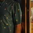 Load image into Gallery viewer, Midsection view of a male model wearing the 7-Strong "Straight Sippin'" Short Sleeve button down. The shirt is a deep green with black ripples as its background, while the foreground has various drinking glasses with various levels of whiskey, some with ice, others without. The model is shown against a background of various whiskies and bourbons on a shelf.