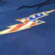 Load image into Gallery viewer, Close up chest region view of the navy blue Freedom Bolt hoodie, showcasing a gold outlined 7-bolt with white stars on blue on top and red and white stripes on the bottom.