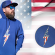 Load image into Gallery viewer, Full view of the navy blue Freedom Bolt hoodie, featured on a male model also sporting the 7-Strong trucker hat and showcasing a gold outlined 7-bolt with white stars on blue on top and red and white stripes on the bottom. The hoodie is featured on an American flag background. There is a detail circle in the bottom right circle featuring the 7-Bolt on the hoodie
