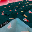 Load image into Gallery viewer, Angled sweep tag view of the 7-Strong "Oh, Christmas Treat" shirt - a green, christmas sweater-like background with columns of various white Christmas tree shaped cakes with red garland, some whole, some bitten. The shirt is displayed against a red, snowing background with a similar parted cake.