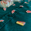 Load image into Gallery viewer, Angled view of the mid-button region on the 7-Strong "Oh, Christmas Treat" shirt - a green, christmas sweater-like background with columns of various white Christmas tree shaped cakes with red garland, some whole, some bitten.