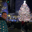 Load image into Gallery viewer, Male model in front of Christmas Tree in a city setting, smiling back to camera sporting the long-sleeve "Oh, Christmas Treat" shirt - a green, Christmas sweater-like background with columns of various white Christmas tree shaped cakes with red garland, some whole, some bitten.