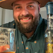 Load image into Gallery viewer, Male model, drinking a bourbon, smiling at camera and wearing the 7-Strong "Straight Sippin'" long Sleeve button down. The shirt is a deep green with black ripples as its background, while the foreground has various drinking glasses with various levels of whiskey, some with ice, others without.