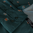 Load image into Gallery viewer, Sleeve and sweep tag view of the 7-Strong "Straight Sippin'" long Sleeve button down. The shirt is a deep green with black ripples as its background, while the foreground has various drinking glasses with various levels of whiskey, some with ice, others without.