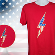 Load image into Gallery viewer, Full view of the red Freedom Bolt unisex tee, featuring a large center placed 7-bolt, outlined in gold, with white stars on blue on top and red and white stripes on the bottom. The shirt is featured on a mannequin against a U.S. flag background. In the bottom left corner, there is a detail circle featuring an up close view of the Freedom Bolt.