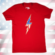 Load image into Gallery viewer, Full centered view of the red Freedom Bolt unisex tee, featuring a large center placed 7-bolt, outlined in gold, with white stars on blue on top and red and white stripes on the bottom. The shirt is featured on a U.S. flag background.