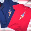 Load image into Gallery viewer, Full view of the red Freedom Bolt unisex tee and the navy Freedom Bolt hoodie overlapping each other, featuring a large center placed 7-bolt, outlined in gold, with white stars on blue on top and red and white stripes on the bottom. Also featured is the 7-Strong trucker hat as a matching comparison. The shirt is featured on a U.S. flag background.