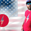 Load image into Gallery viewer, Full view of the red Freedom Bolt unisex tee, featuring a large center placed 7-bolt, outlined in gold, with white stars on blue on top and red and white stripes on the bottom. The shirt is featured on a medium male model against a U.S. flag background. In the bottom left corner, there is a detail circle featuring an up close view of the Freedom Bolt.