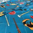 Load image into Gallery viewer, Middle buttons view of the 7-Strong "TLC OMG" shirt, a homage to wrestling objects featured all over the shirt including tables, ladders, chairs, ring bells, sledgehammers, brass knuckles, championships, guitars, stop signs, microphones amongst other items. The items are on a blue shirt with a faded steel cage background. The shirt is featured on a background of a wrestling ring with a standing ladder.