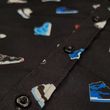 Load image into Gallery viewer, Midsection close up view of the youth short sleeve version of "Got 'Em!" The button down features various athletic fashion sneakers in an 80bit format arranged all over a black shirt. The shirt is featured against a background of a basketball player jumping in air, noticeable sneakers being worn.