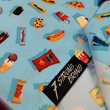 Load image into Gallery viewer, Bottom sweep tag view of the 7-Strong "Cheat Day" youth button down featuring various snacks, treats, and fast food features on a light blue shirt. The shirt is displayed against a background of a gym weight rack