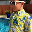 Load image into Gallery viewer, Youth model wearing the 7-Strong "Late Bloomer" youth button down - a yellow background shirt with white and blue flowers patterned all over it. Model is standing by the pool in sunglasses with the shirt being shown at a medium close up on the body.