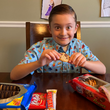 Load image into Gallery viewer, Child model wearing the 7-Strong "Cheat Day" youth button down featuring various snacks, treats, and fast food features on a light blue shirt indulging in some cookies and candy.