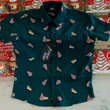 Load image into Gallery viewer, Full view of the 7-Strong "Oh, Christmas Treat" shirt - a green, christmas sweater-like background with columns of various white Christmas tree shaped cakes with red garland, some whole, some bitten. Shirt dlaided out on a display of tree cake boxes.