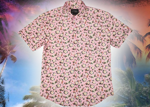 Full centered view of the "Magnolia PI" 7-Strong Adult button down shirt, featuring white and green magnolia flowers patterned all over a light pink background. The shirt itself is displayed against a palm tree tropical background.