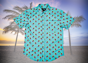 Full centered view of the adult 7-Strong "Sunset Palms" shirt, featuring small yellow and pink suns, reminiscent of a sunset, behind a silhouette of a palm tree and birds, patterned at various angles all over the turquoise-like shirt. The shirt is featured against a photo of a palm tree beach at sunset.