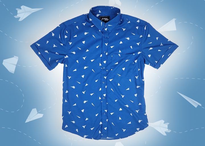 A complete view of the adult 7-Strong "Paper Planes" button down shirt - featuring various sizes and designs of paper airplanes, some with dotted trails behind them against a royal blue background. The shirt is displayed against a faced background of the shirt details.