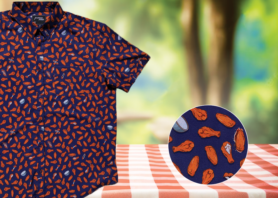 3/4 view of the 7-Strong "Wingman" shirt, featuring various different flat and drum chicken wings and dip sauces on a deep blue background. Shirt is featured against a picnic/park background. Bottom right corner features a detail circle showing the design in close up. 