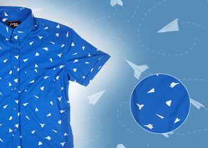 A complete view of the adult 7-Strong "Paper Planes" button down shirt - featuring various sizes and designs of paper airplanes, some with dotted trails behind them against a royal blue background. The shirt is displayed against a faced background of the shirt details. Bottom right features a detail circle highlighting the shirt design. 