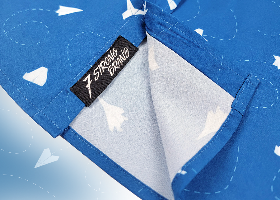 A sweep tag view of the adult 7-Strong "Paper Planes" button down shirt - featuring various sizes and designs of paper airplanes, some with dotted trails behind them against a royal blue background. The shirt is displayed against a faced background of the shirt details.