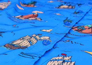 Close up, middle button view of the 7-Strong "7-Seas" shirt, a bright blue colored shirt with various nautical depictions such as islands, ships, mermaids, etc - drawn in a treasure map like fashion.