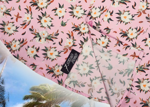 Bottom sweep tag close up view of the "Magnolia PI" 7-Strong Adult button down shirt, featuring white and green magnolia flowers patterned all over a light pink background. The shirt itself is displayed against a palm tree tropical background.