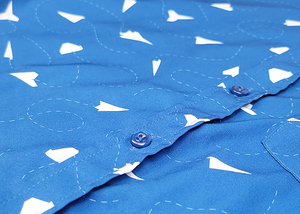 A midsection button view of the adult 7-Strong "Paper Planes" button down shirt - featuring various sizes and designs of paper airplanes, some with dotted trails behind them against a royal blue background. The shirt is displayed against a faced background of the shirt details.