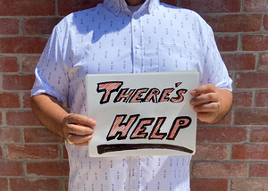 A midsection view of a male model wearing the 7-Strong "Stay" button down, a white shirt with staggered, hollowed out semicolons outlined in teal and purple - the colors for suicide awareness. Model is holding a handwritten sign saying "There's Help."