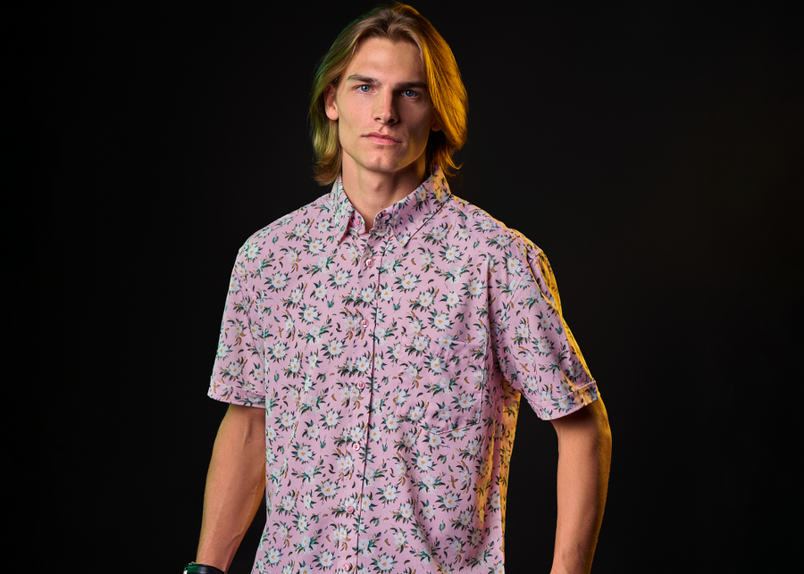 Medium shot of a male model sporting the "Magnolia PI" 7-Strong Adult button down shirt, featuring white and green magnolia flowers patterned all over a light pink background. 