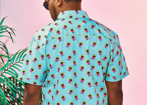 Back, medium view of male model wearing the adult 7-Strong "Sunset Palms" shirt, featuring small yellow and pink suns, reminiscent of a sunset, behind a silhouette of a palm tree and birds, patterned at various angles all over the turquoise-like shirt. The model is featured against a pink tropical backdrop with palm trees in the background. 