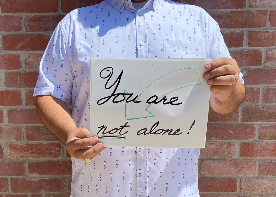 A midsection view of a male model wearing the 7-Strong "Stay" button down, a white shirt with staggered, hollowed out semicolons outlined in teal and purple - the colors for suicide awareness. Model is holding a handwritten sign saying "You Are Not Alone."