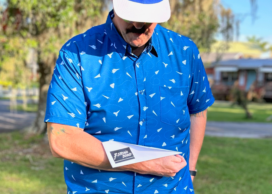 Male model wearing the adult 7-Strong "Paper Planes" button down shirt - featuring various sizes and designs of paper airplanes, some with dotted trails behind them against a royal blue background. Model is looking down admiring his paper airplane which displays the 7-Strong Brand logo.