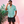 Load image into Gallery viewer, Full view of  male model wearing the adult 7-Strong &quot;Sunset Palms&quot; shirt, featuring small yellow and pink suns, reminiscent of a sunset, behind a silhouette of a palm tree and birds, patterned at various angles all over the turquoise-like shirt. The model is featured against a pink tropical backdrop with palm trees in the background.

