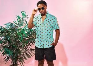 Full view of  male model wearing the adult 7-Strong "Sunset Palms" shirt, featuring small yellow and pink suns, reminiscent of a sunset, behind a silhouette of a palm tree and birds, patterned at various angles all over the turquoise-like shirt. The model is featured against a pink tropical backdrop with palm trees in the background.