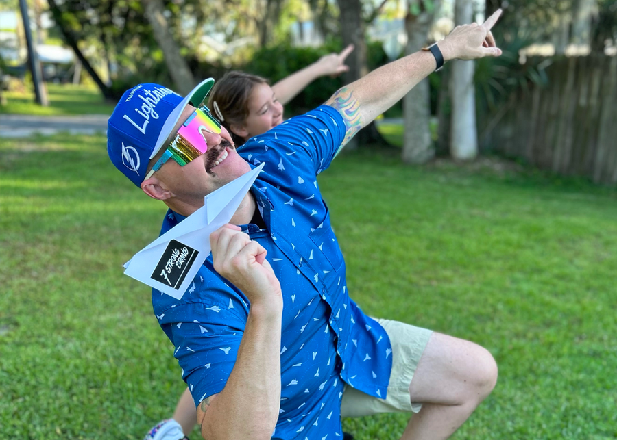 Male model and female child showcasing the adult 7-Strong "Paper Planes" button down shirt - featuring various sizes and designs of paper airplanes, some with dotted trails behind them against a royal blue background. Model is pointing towards skill, smiling at camera, as if aiming the paper airplane in is hand towards a location. 