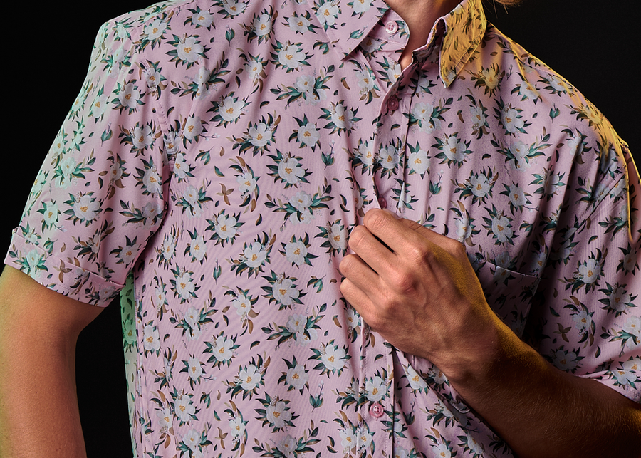 Medium Close Up view a male model, tugging on the fabric, wearing the "Magnolia PI" 7-Strong Adult button down shirt, featuring white and green magnolia flowers patterned all over a light pink background. 