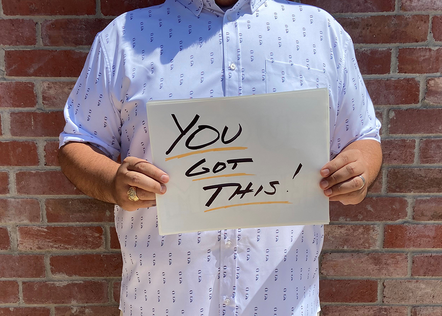 A midsection view of a male model wearing the 7-Strong "Stay" button down, a white shirt with staggered, hollowed out semicolons outlined in teal and purple - the colors for suicide awareness. Model is holding a handwritten sign saying "You Got This!"