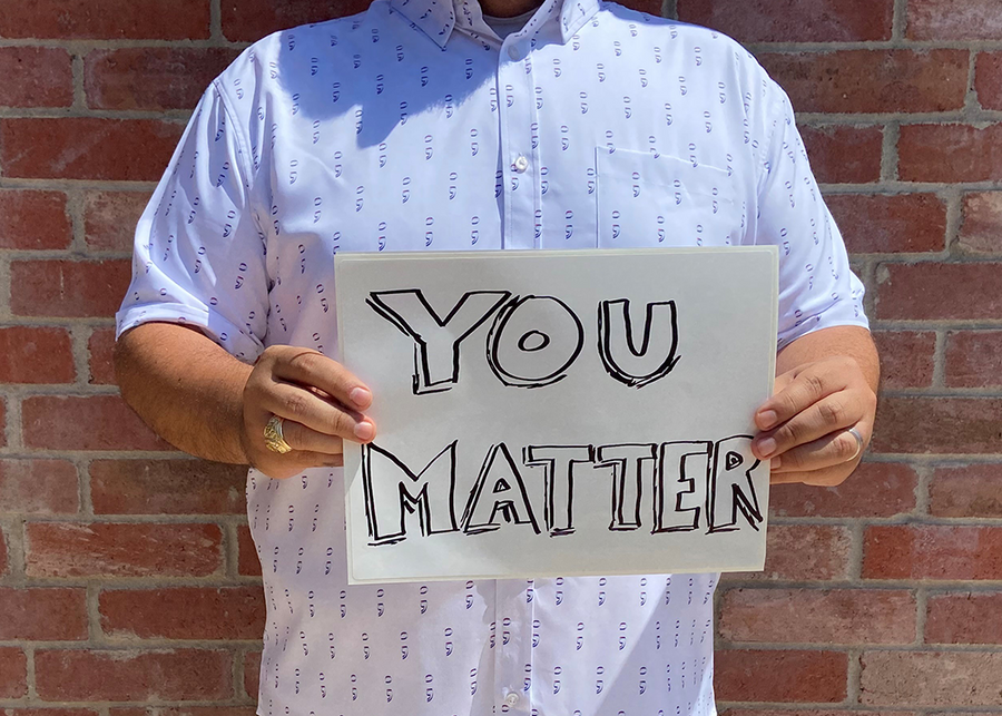 A midsection view of a male model wearing the 7-Strong "Stay" button down, a white shirt with staggered, hollowed out semicolons outlined in teal and purple - the colors for suicide awareness. Model is holding a handwritten sign saying "You Matter."